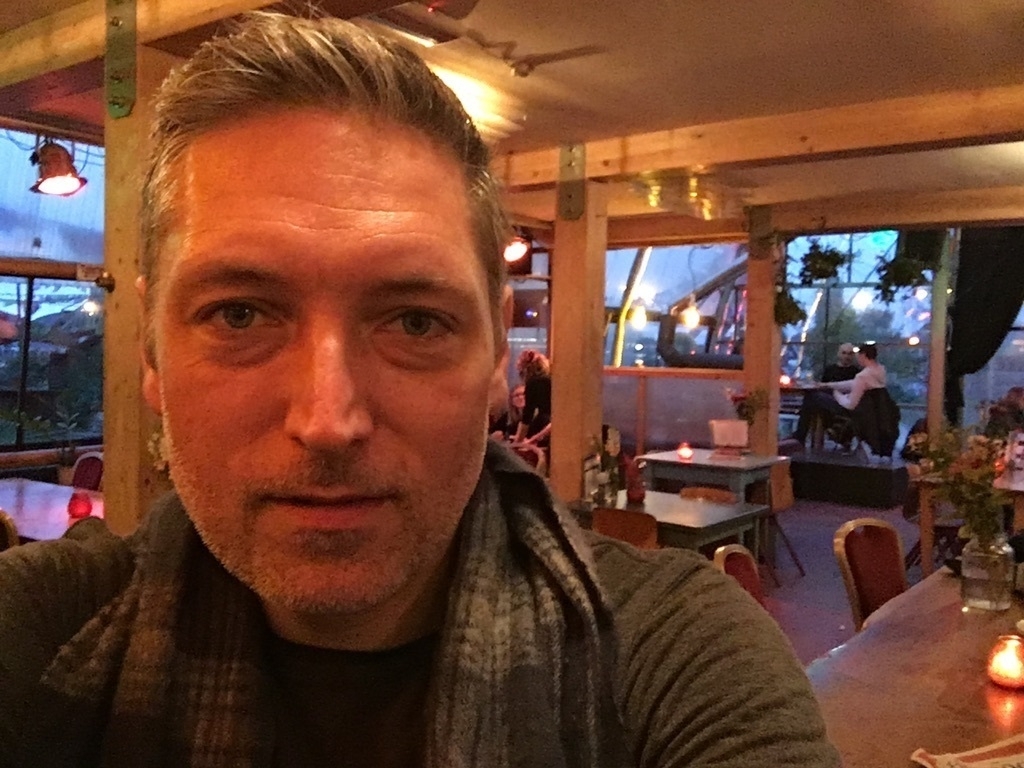  Selfie against a backdrop of a well-lit bar with many windows and flowers adorning tables. I'm looking right into the camera with wind-swept hair and a scarf.